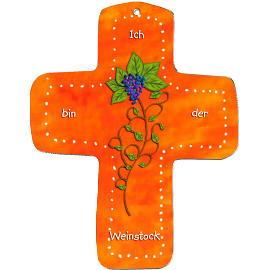 Das metALUm Wandkreuz aus orangefarbenem MDF hat einen gepunkteten weißen Rand, eine gemalte Weinrebe mit grünen Blättern und lila Trauben und den Text "Ich bin der Weinstock". Dank des Lochs an der Spitze eignet es sich perfekt als Geschenk für Kinder.